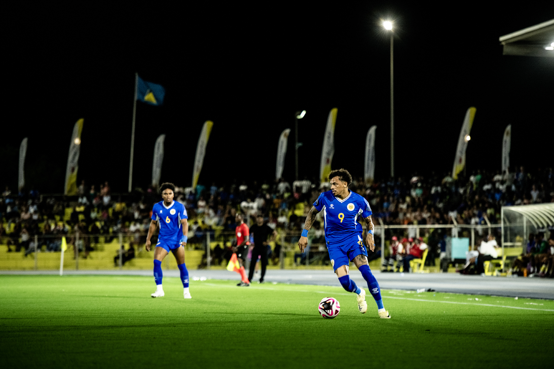 Soccer players in blue uniforms competing on a lit stadium field at night, with a large crowd in the stands.