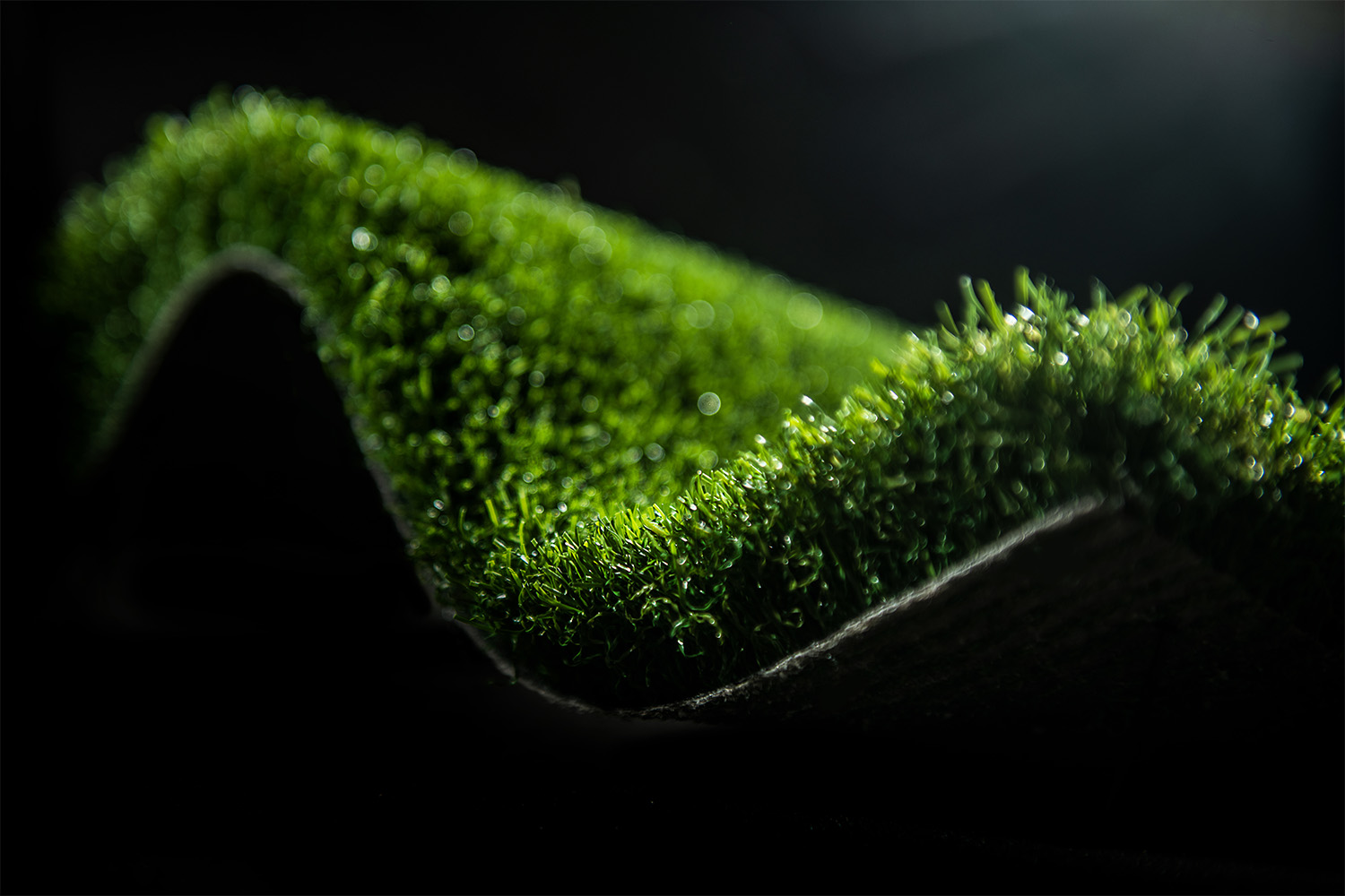 Angled close-up of a rolled piece of artificial turf (GreenFields Pure PT), showing dense green grass fibers highlighted by strong contrast lighting to emphasize texture and depth
